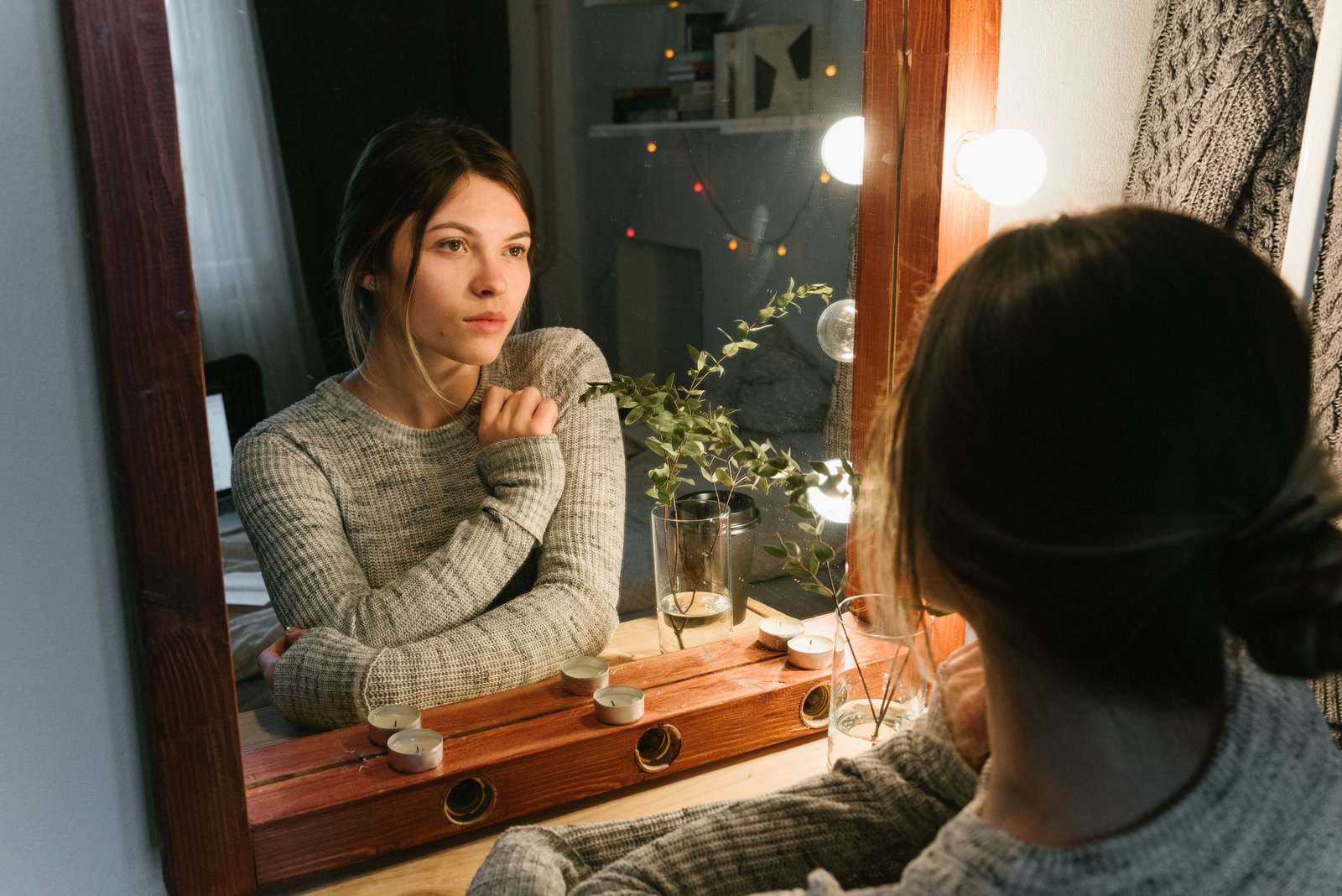 A woman in a cozy sweater contemplates her reflection in a warmly lit room.